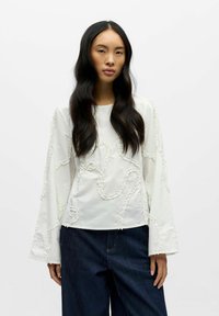 Hvit langermet skjorte med teksturerte volangdetaljer i et bølgemønster, kombinert med mørk denim bukser med vide ben.