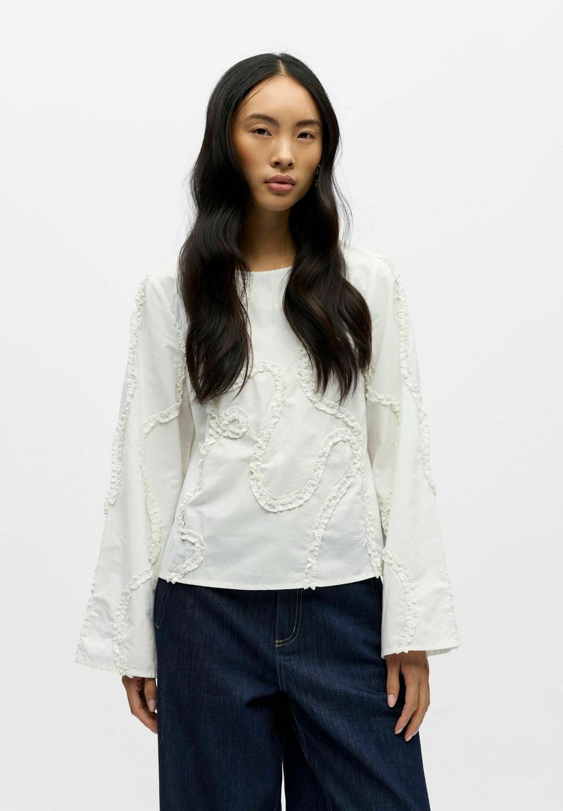 Hvit langermet skjorte med teksturerte volangdetaljer i et bølgemønster, kombinert med mørk denim bukser med vide ben.