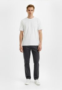 White cotton t-shirt with short sleeves, paired with dark gray trousers and white sneakers. Model stands against a neutral background.