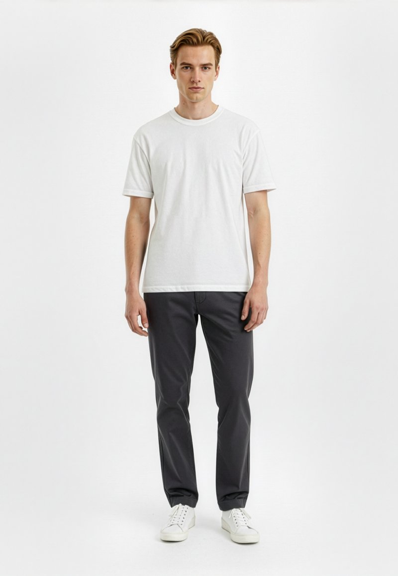 White cotton t-shirt with short sleeves, paired with dark gray trousers and white sneakers. Model stands against a neutral background.