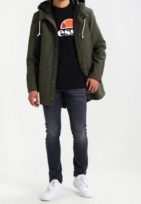 Olivgrön ytterjacka med huva, svart innertyg, framknappar, bärs över en svart sweatshirt med vitt logotyp. Matchas med mörka jeans och vita sneakers.