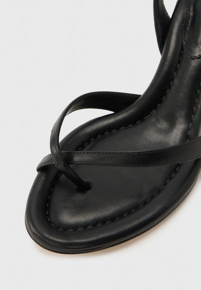 Μαύρο δερμάτινο σανδάλι με λεπτό λουράκι στα δάχτυλα και λουράκι που διασταυρώνεται κοντά στο εμπρόσθιο μέρος της σόλας, με ορατή ραφή κατά μήκος των ακμών.