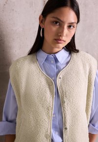 Gilet en polaire écru avec une finition texturée, fermetures à pression, porté par-dessus une chemise à boutons rayée bleu clair avec les manches retroussées.