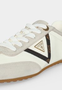 Baskets Guess blanches et beiges avec lacets blancs, accents en daim beige, bordure noire à motifs et logo triangulaire Guess sur le côté.