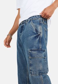 Pantaloni cargo in denim blu con una vestibilità rilassata. Presentano una tasca laterale, cuciture visibili e una finitura sbiadita. Indossati con una camicia bianca.
