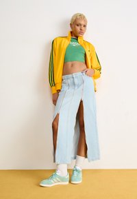 Chaqueta amarilla con cremallera y rayas verdes, top corto verde, falda larga de mezclilla azul claro con cremalleras laterales y zapatillas verde menta.