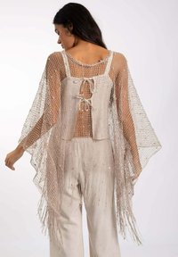 Poncho de malla beige con la espalda descubierta y detalle de lazo, con flecos a lo largo del dobladillo y mangas sueltas. Combinado con pantalones de color claro.