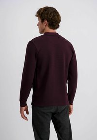 Un jeune homme de dos portant un polo à manches longues de couleur bordeaux foncé et un pantalon à carreaux sombres devant un fond uni.