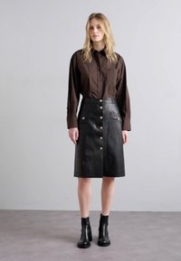 Zwarte leren rok met gouden knopen, twee voorzakken en een rechte snit, gecombineerd met een donkerbruine blouse met lange mouwen en enkellaarsjes.