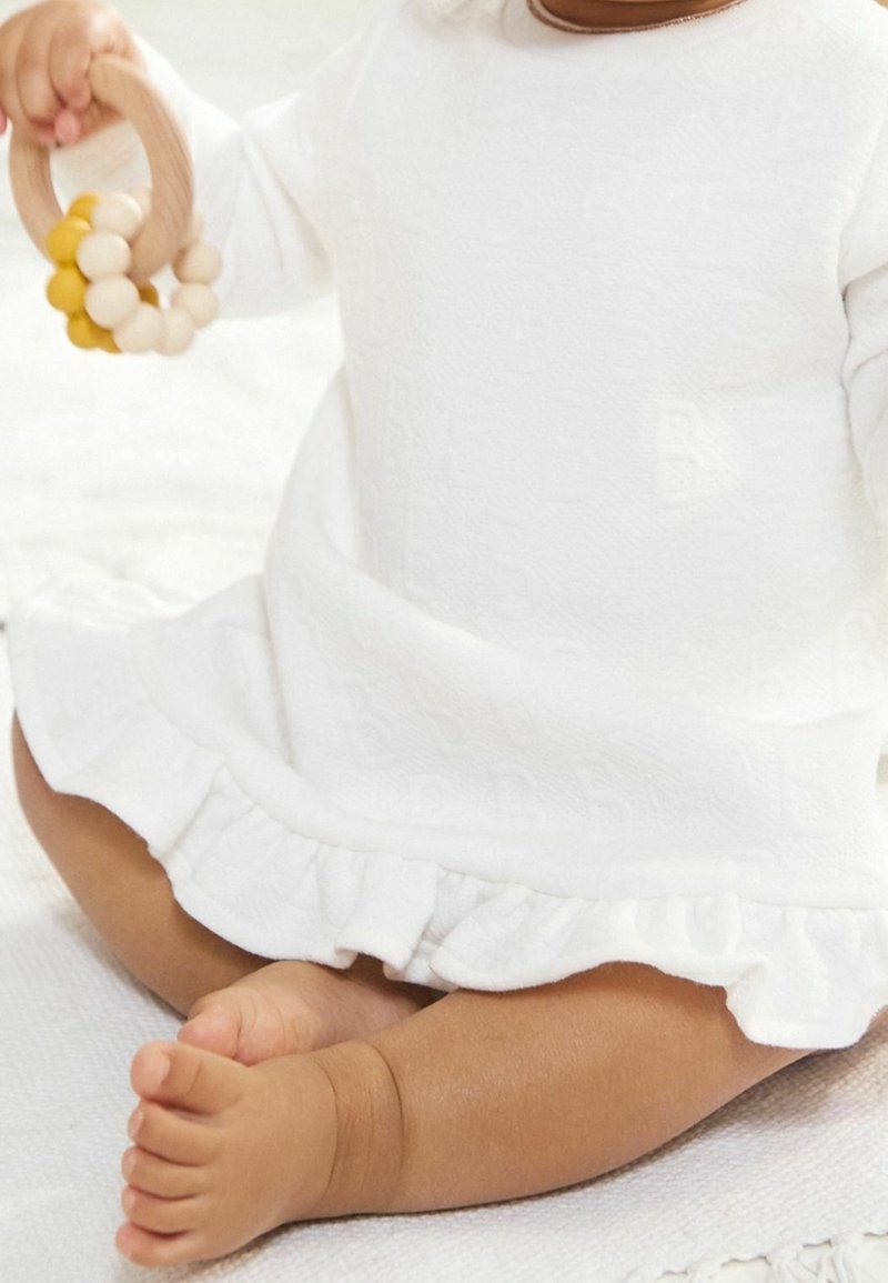 Bébé assis sur une couverture blanche, portant une robe blanche à volants, tenant un jouet anneau en bois avec des perles beige et jaune.