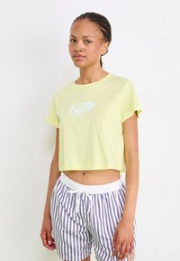 Kortermet, cropped t-skjorte i lys gul bomull med hvit blomstergrafikk. Bært med hvite stripete shorts med snor i livet.