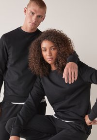 Czarne bawełniane bluzy i dresy, z długimi rękawami i ściągaczami, noszone przez dwóch modeli w neutralnym otoczeniu.