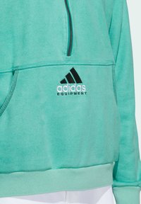 Tyrkysový fleecový pulover s vysokým límcem, zipem vpředu a černým logem Adidas. Má žebrované manžety a lem pasu. Hladká textura.