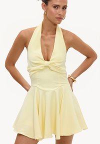 Femme portant une robe jaune pâle à col licou avec un détail nœud à la taille, accessoirisée avec des boucles d'oreilles créoles en or et un bracelet en or.