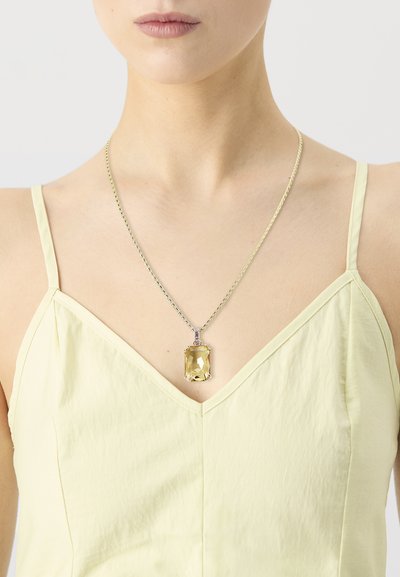 Femme portant un top à fines bretelles jaune pâle avec un collier en argent mettant en vedette un grand pendentif en forme de rectangle en pierre précieuse jaune.