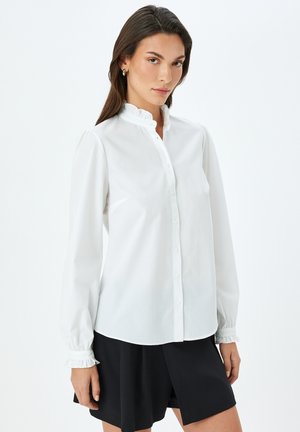 Blusa blanca con un alto cuello con volantes, mangas largas y un cierre frontal de botones. Presenta detalles fruncidos en los puños de las mangas. Diseño simple.