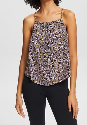 Kobieta w bezrękawniku w kwiatowy wzór z cienkimi ramiączkami i czarnych dopasowanych spodniach, stojąca na jednolitym tle.