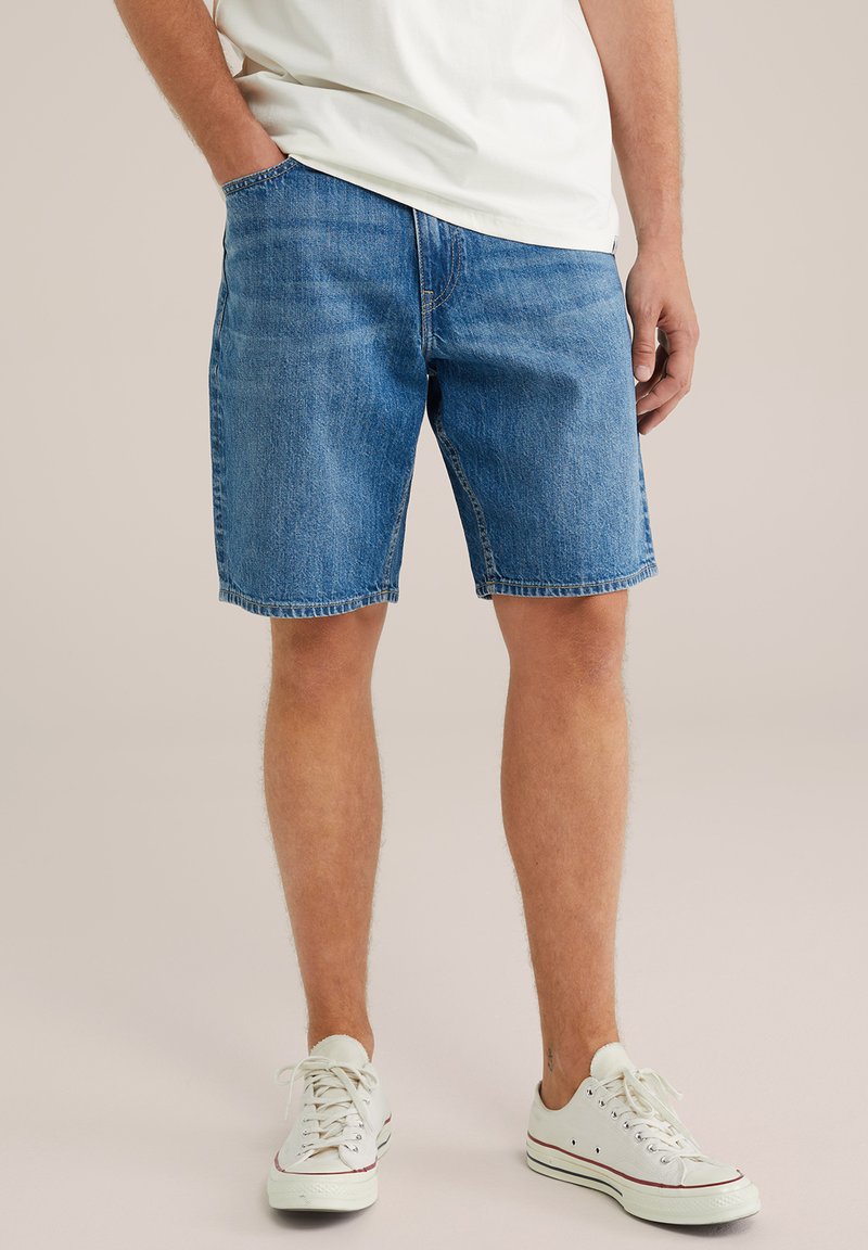 Traper šorts u srednje plavoj boji s opuštenim krojem, s pet džepova i pojačanim šavovima. Upareni s bijelim platnenim tenisicama.