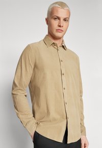 Chemise en coton beige à boutons, avec un col, des manches longues et une coupe décontractée. Présente des boutons marron et un tissu texturé.