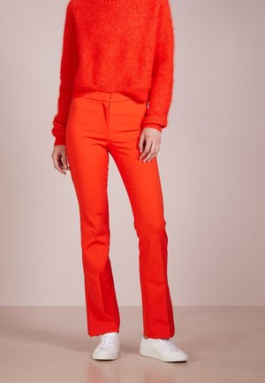 Personne portant un pantalon rouge vif et un pull rouge assorti avec des baskets blanches, debout devant un fond uni.