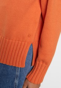 Maglione arancione a maglia con orlo e polsini a coste, caratterizzato da fessure laterali. Texture morbida e un piccolo, sottile dettaglio del logo vicino all'orlo.