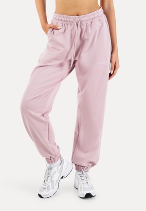 Frau trägt hellrosa Jogginghose mit elastischem Bund und weiße Sportschuhe, steht mit einer Hand in der Tasche und überkreuzten Beinen.