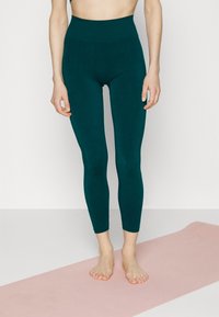 Leggings taille haute teal en tissu lisse et extensible. Ils s'ajustent parfaitement et descendent jusqu'aux chevilles, accompagnés d'un tapis de yoga rose clair.