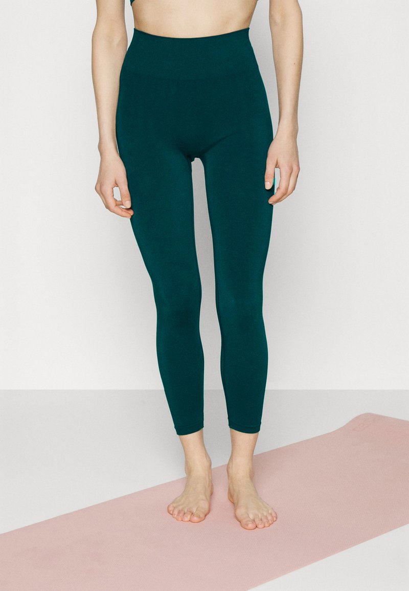Leggings taille haute teal en tissu lisse et extensible. Ils s'ajustent parfaitement et descendent jusqu'aux chevilles, accompagnés d'un tapis de yoga rose clair.