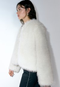 Jachetă albă din blană artificială cu design scurt, textură pufoasă proeminentă și mâneci largi, asortată cu pantaloni negri mulați cu ținte metalice.