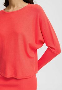 Pull en tricot corail avec une coupe ample, un col rond et des coutures d'épaule tombantes. Texture douce avec des accents côtelés à l'ourlet et aux poignets.