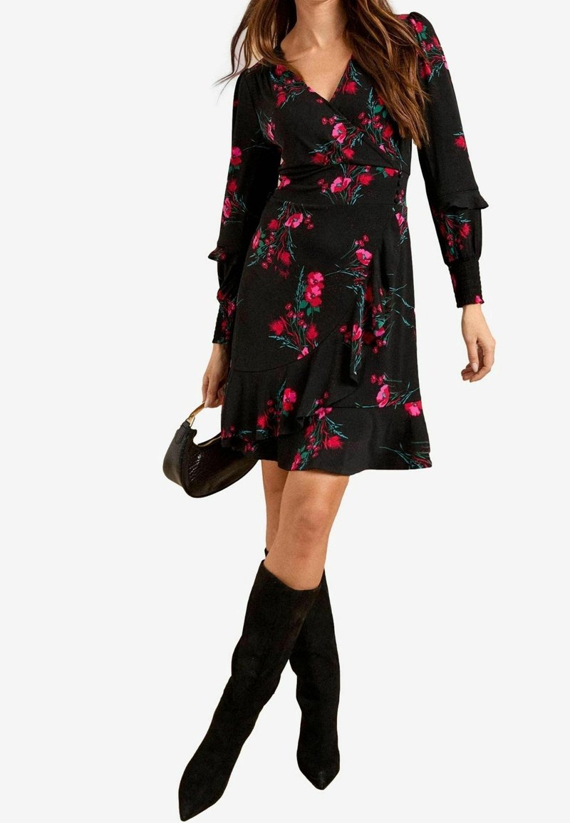 Femme portant une robe portefeuille noire à fleurs avec des manches longues, tenant un petit sac à main noir et portant des bottes noires mi-mollet.