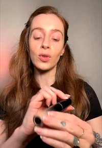 Frau mit langen braunen Haaren hält und öffnet eine schwarze Lippenstift-Tube vor einem schlichten hellen Hintergrund.
