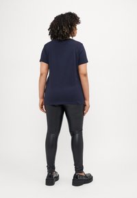 Tumši zila īsām piedurknēm t-krekls, kas apvienots ar spīdīgiem melniem leggins un platformas apaviem ar uzspiesta aksesuāriem, skatīts no mugurpuses.