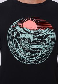 Zwarte katoenen t-shirt met een grafisch ontwerp van gestileerde golven en bergen in mintgroen, met een roze zon en lijnen die naar buiten uitbreiden.