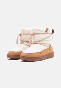 Valkoiset pörröiset nilkkurit, joissa on tan-nahkaiset yksityiskohdat, säädettävät beige-nauhat ja teksturoitu kumipohja. Logo näkyvissä sivulla ja kannassa.