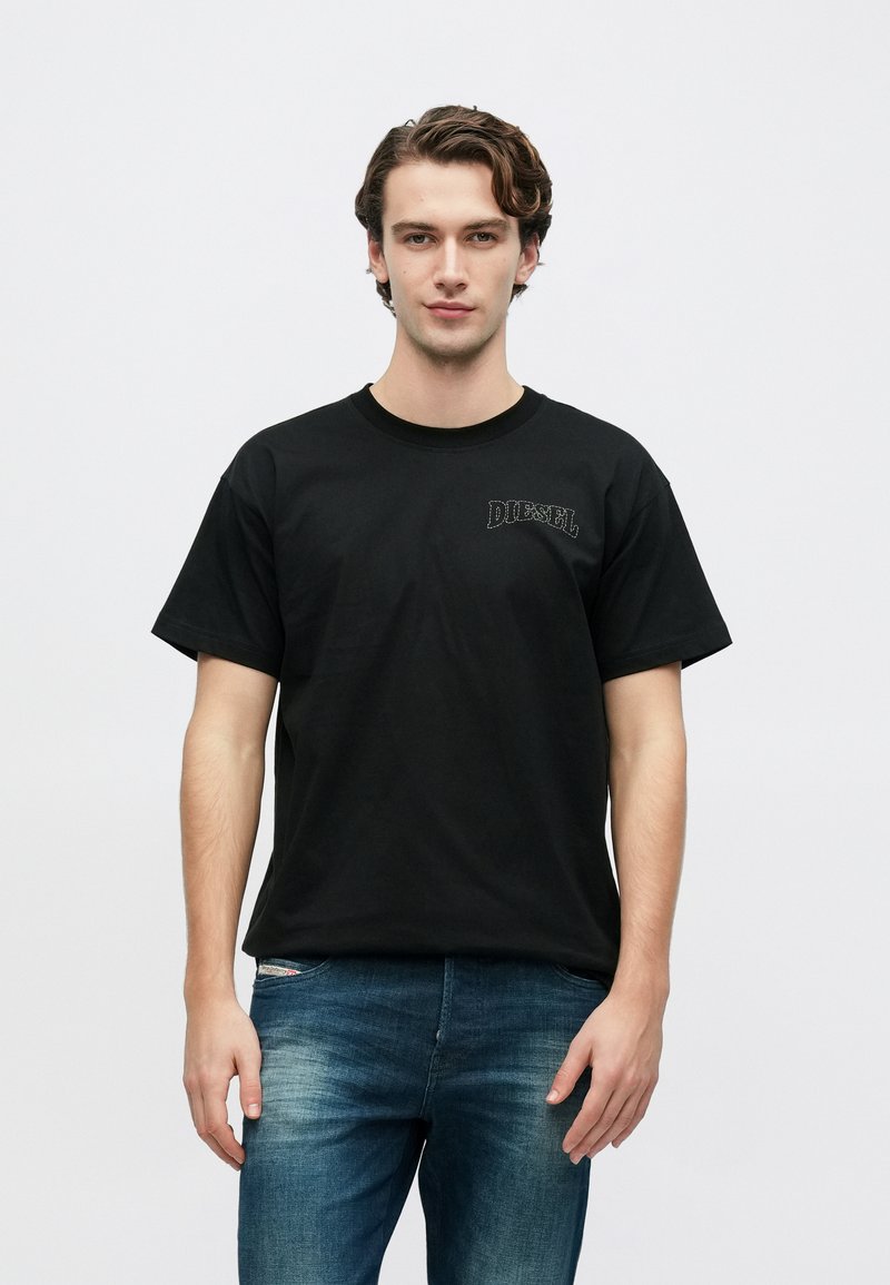 Μαύρο βαμβακερό t-shirt με κοντά μανίκια, στρογγυλή λαιμόκοψη και διακριτικό λογότυπο "DIESEL" στο μπροστινό μέρος. Φορεμένο με μπλε ξεβαμμένο τζιν παντελόνι.