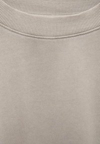 Svijetlosmeđa dukserica s rebrastim izrezom. Tekstura je glatka i mekana. Šavovi su vidljivi, što pruža strukturni detalj.