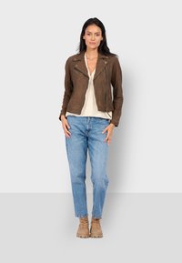 Hnědá semišová motorkářská bunda se zipovými kapsami, v kombinaci se světle modrými džíny a tanovými šněrovacími botami, na neutrálním pozadí.