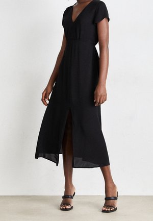 Kobieta w czarnej sukience midi z dekoltem w serek, krótkim rękawem i przednim rozcięciem, w połączeniu z czarnymi sandałami na obcasie z odkrytymi palcami na neutralnym tle.