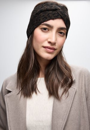 Fascia per testa lavorata a maglia nera e marrone con design a torsione frontale, tessuto testurizzato, indossata con un maglione e un cappotto di colore chiaro.