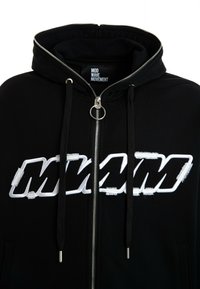 Sudadera negra con cremallera y un gran logo "MMM" desgastado en blanco. Cuenta con cordones, una cremallera plateada y bolsillos laterales. Textura de tela suave.