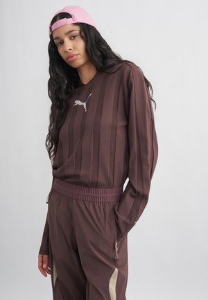 Femme aux longs cheveux foncés portant une casquette rose à l’envers, un sweat-shirt rayé marron Puma et un pantalon assorti marron avec des accents beiges.