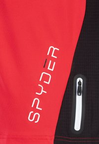 Chaqueta deportiva roja y negra con un bolsillo con cremallera, tejido texturizado y logo "SPYDER" en blanco en la sección roja.