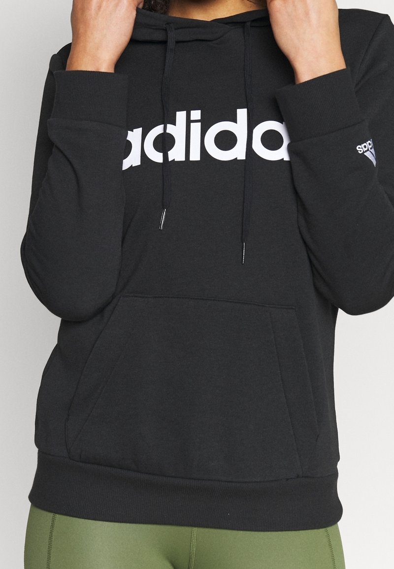 Personne portant un sweat à capuche Adidas noir avec un logo blanc et des leggings verts, mains tenant le sweat près du cou.
