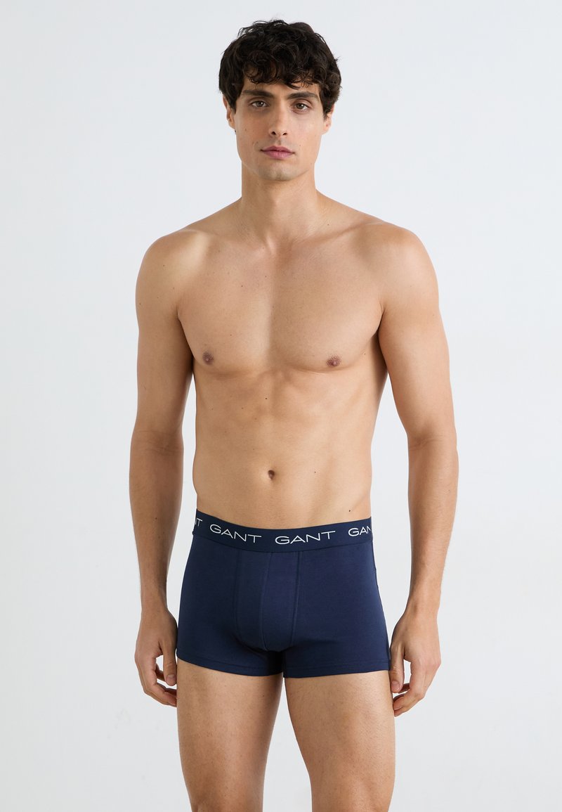 Oberkörperfreier Mann mit lockigem dunklem Haar, der dunkelblaue Boxershorts mit „GANT“-Logo am Bund trägt, steht vor einem schlichten hellen Hintergrund.