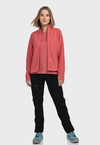 Fleece-Jacke in Koralle, mit durchgehendem Reißverschluss und seitlichen Taschen, kombiniert mit einem Koralle-Oberteil und schwarzen Hosen. Blaue Wanderschuhe runden das Outfit ab.