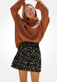 Oversized svetr v rezavé barvě pletený s vysokým límcem, v kombinaci s černou mini sukní s květinovým potiskem a vrstveným designem. Texturované materiály a elastický pas.