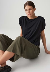 Schwarzes, lässiges T-Shirt mit kurzen Ärmeln, kombiniert mit olivgrünen, weit geschnittenen Hosen. Das Model sitzt vor einem neutralen Hintergrund. Schwarze Schuhe sind sichtbar.