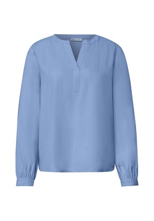 Femme portant un chemisier bleu et un jean à jambes larges, debout les mains dans les poches, portant un sac à main marron, devant un mur clair uni.
