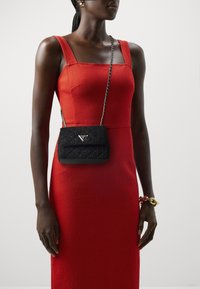 Donna che indossa un vestito rosso aderente senza maniche, con una piccola borsa a tracolla nera e un bracciale spesso oro e rosso sul polso destro.
